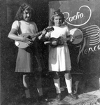 108747 Portret van twee meisjes met mandoline en mini-gitaar staande voor een reclamebord van Radio Henco in de ...